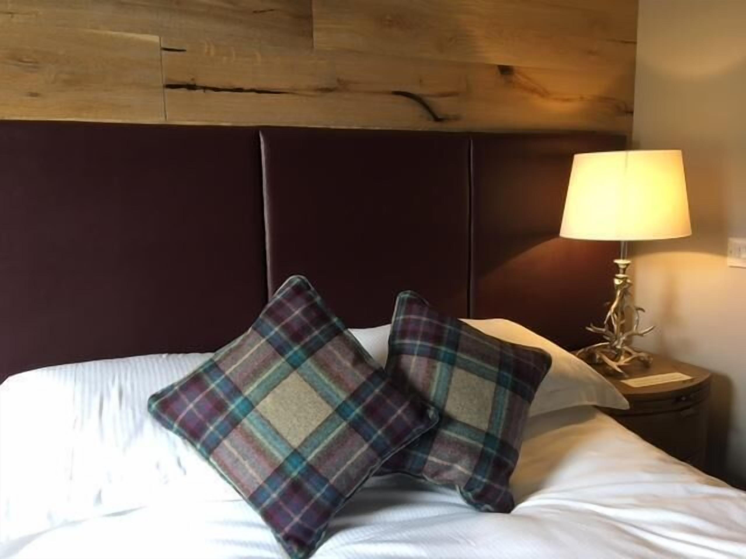 A bed with white bedding, two plaid pillows, a wooden headboard, and a bedside lamp.