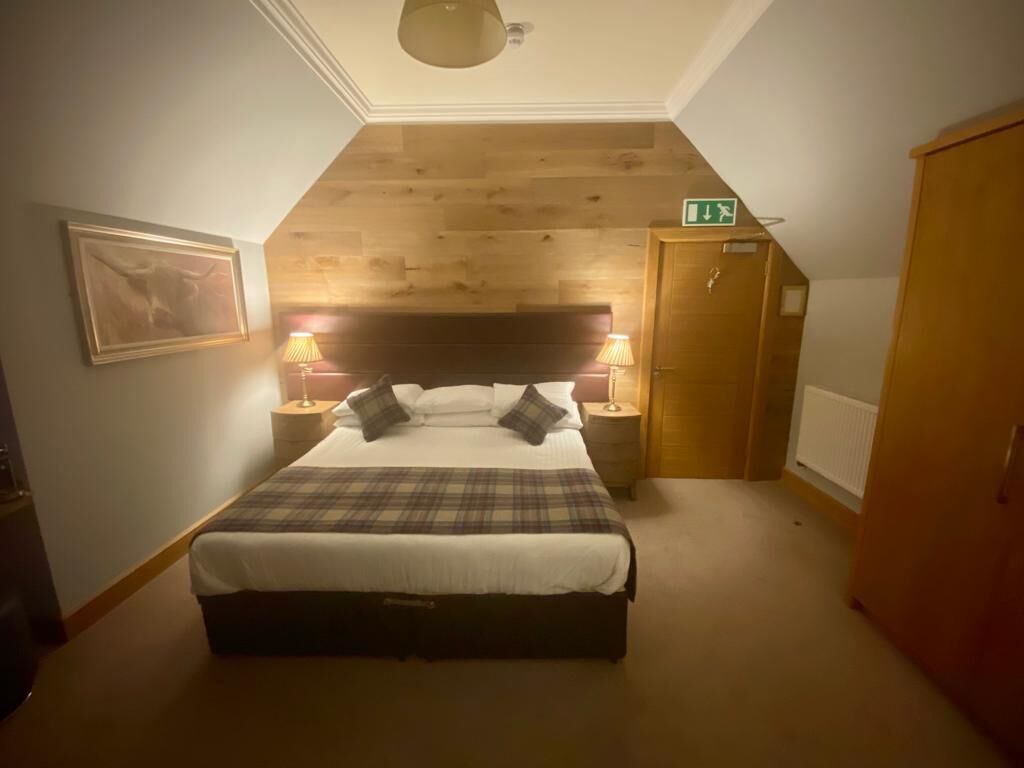 Une chambre avec un plafond en bois, un lit recouvert d’une couverture à carreaux, deux lampes de chevet et un tableau encadré au mur.