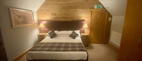 Une chambre avec un plafond en bois, un lit recouvert d’une couverture à carreaux, deux lampes de chevet et un tableau encadré au mur.