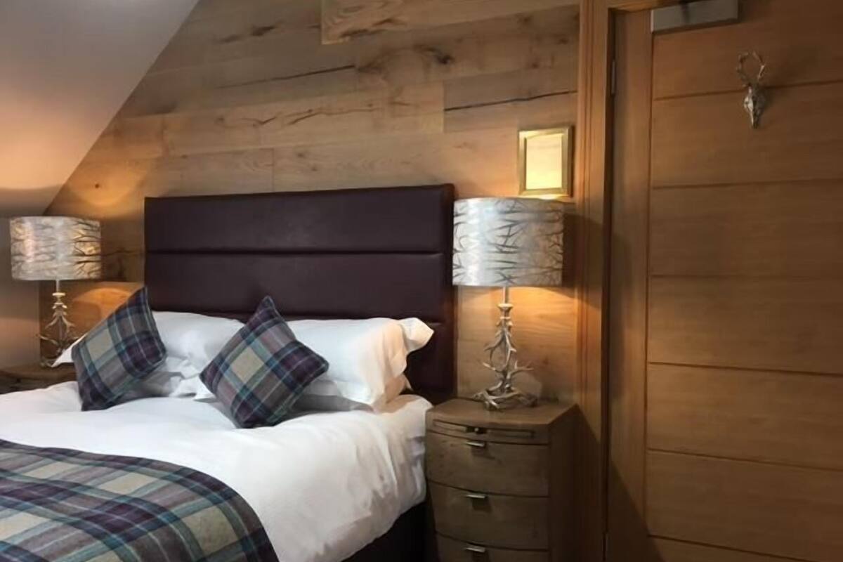 A bedroom with a wooden ceiling, a dark headboard, two bedside lamps, a wooden nightstand, and a wardrobe.