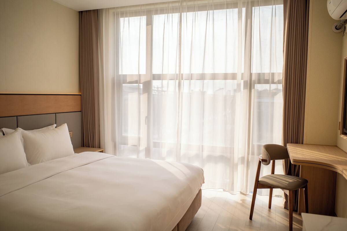Habitación de hotel con una cama grande, un escritorio con una silla y vistas a través de cortinas translúcidas.