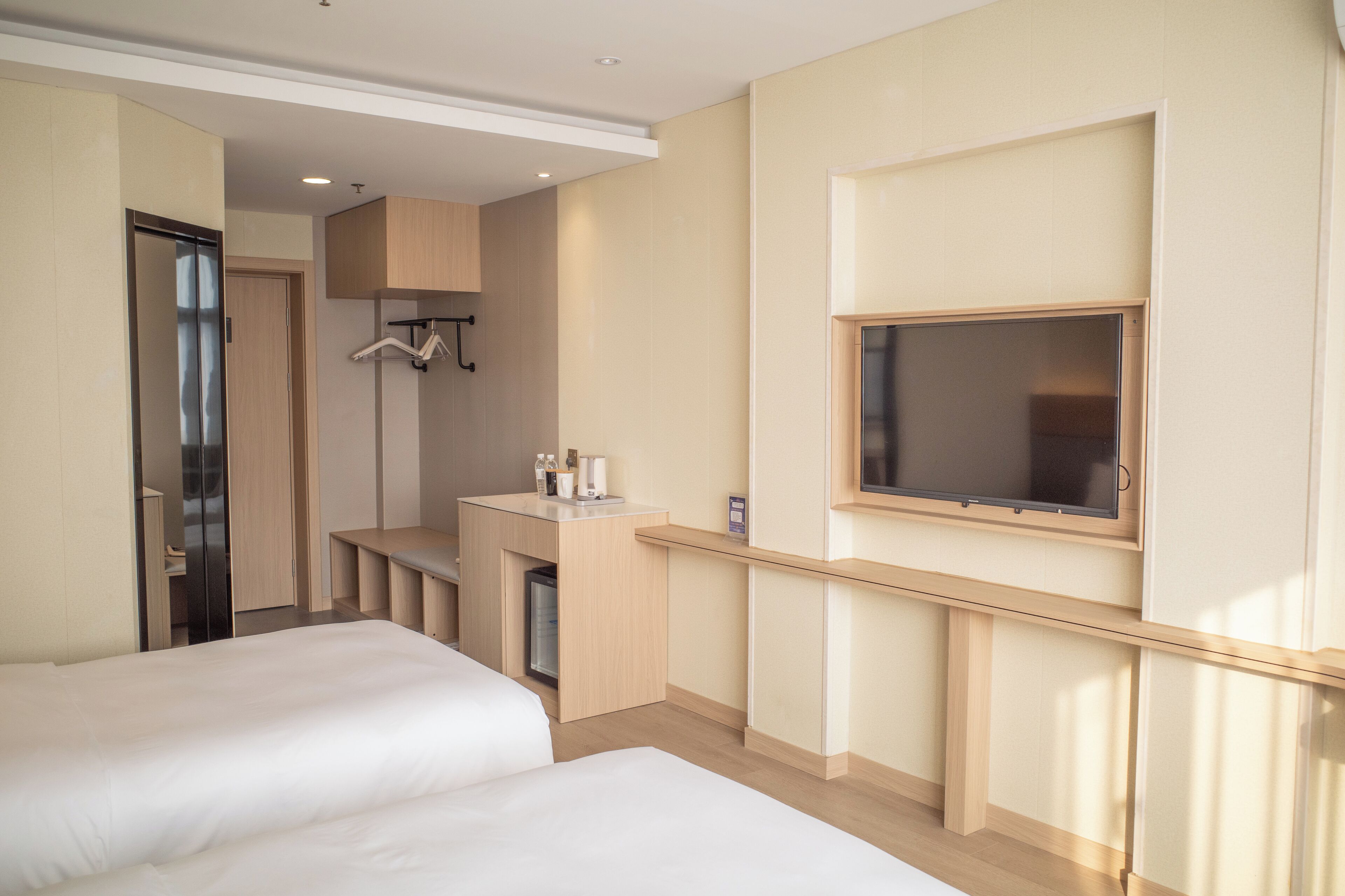 A hotel room with two beds, a small kitchenette, a TV mounted on the wall, and a wardrobe.