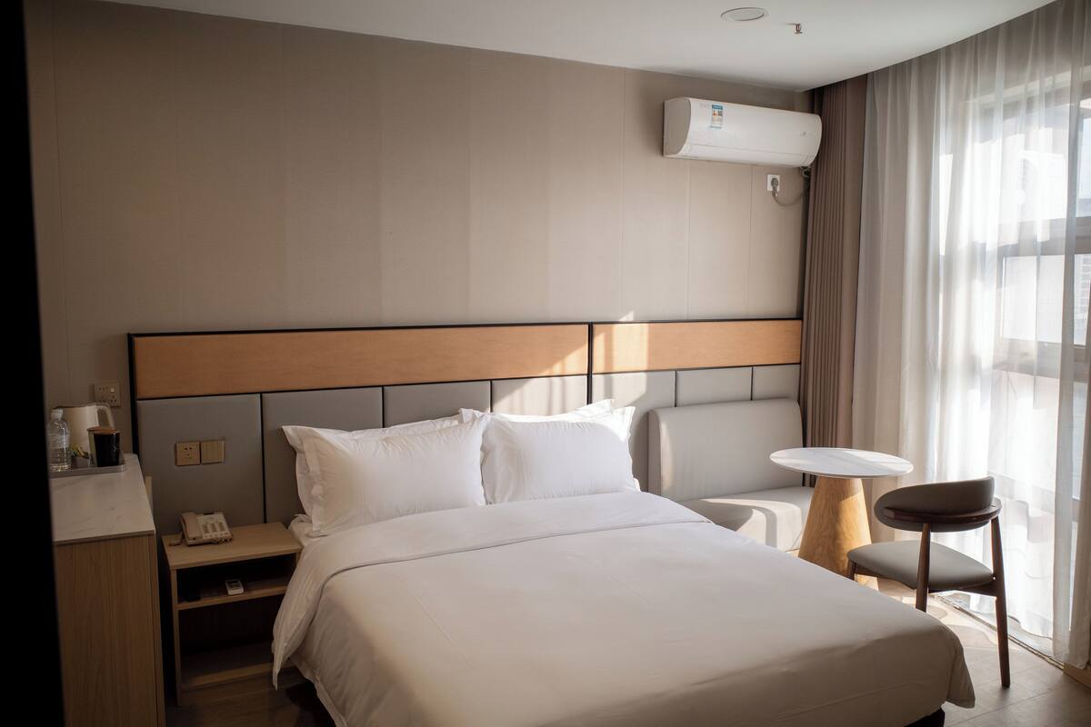 Habitación de hotel con una cama grande, mesitas de noche, una silla, una pequeña mesa redonda y un equipo de aire acondicionado en la pared.