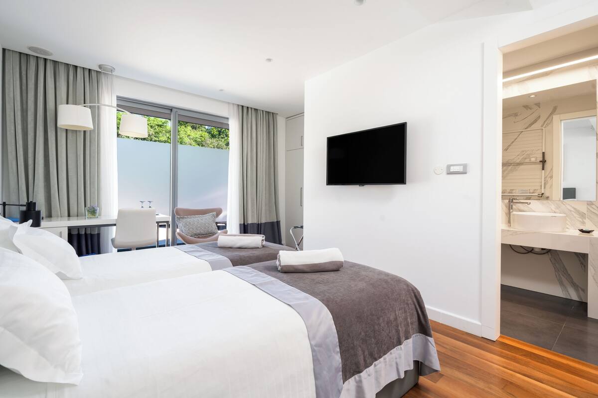 A hotel room with two beds, a TV, a desk, and a bathroom with a mirror and sink.
