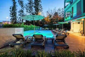 A poolside area with lounge chairs and umbrellas, surrounded by greenery and a modern building.