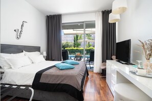 A modern hotel room with a bed, a TV, a desk, and a balcony with a view of buildings and greenery.
