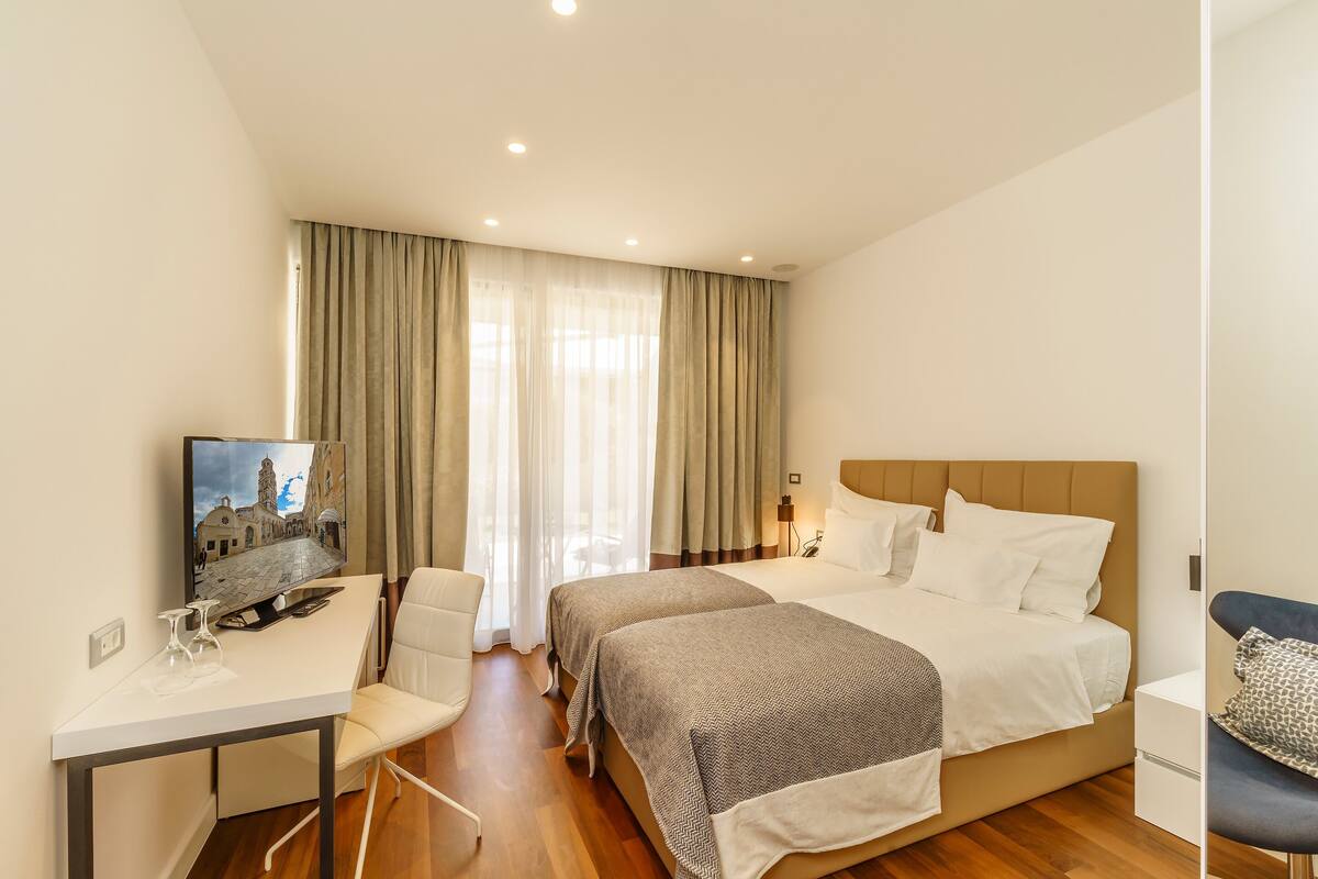 A hotel room with a bed, bedside tables, a desk with a chair, and a large window with curtains.