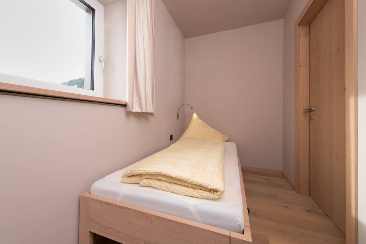 A small, single-bed room with a wooden bed frame, a window with a curtain, and a wooden door.