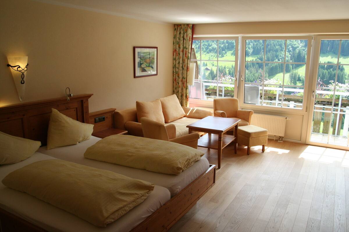 A room with two beds, a sofa, a table, and a chair. There is a window with a view of greenery outside.