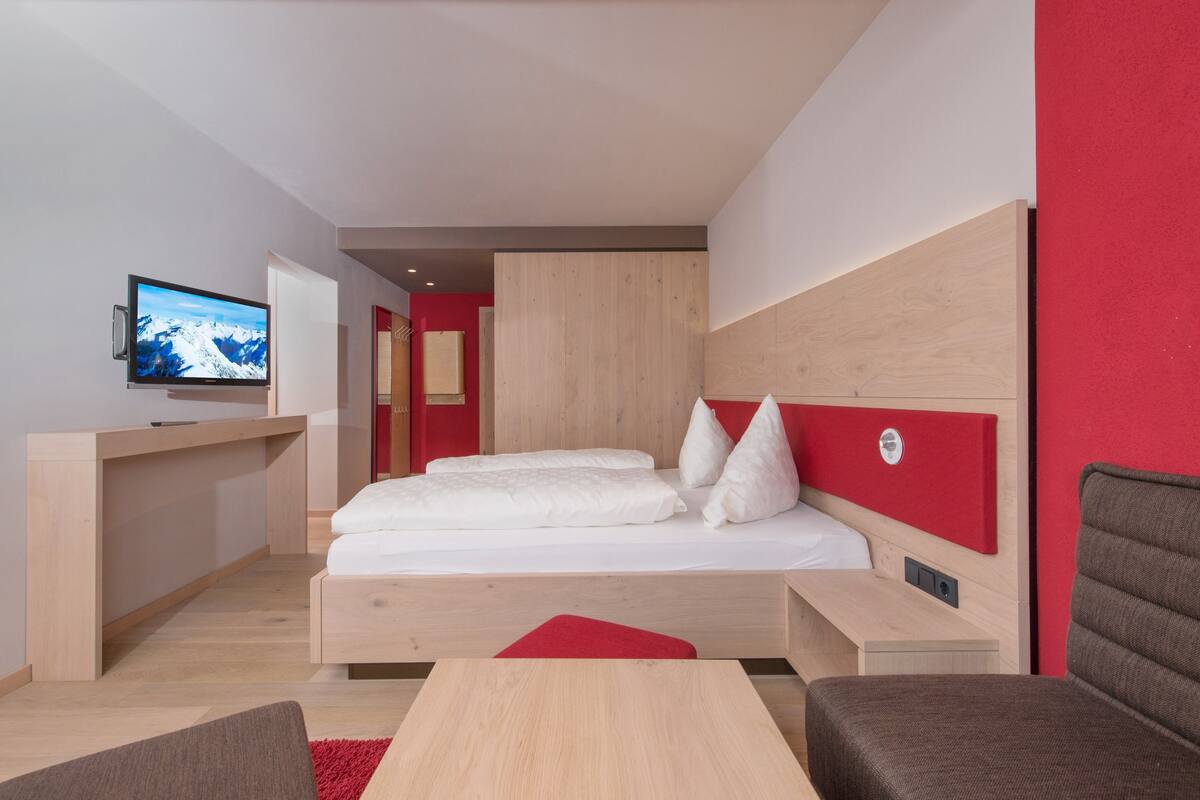A modern hotel room with a bed, a desk, a TV, and a red accent wall.