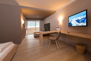 A modern hotel room with a wooden desk, a comfortable chair, a bed, a TV mounted on the wall, and a balcony with a view of snow-capped mountains. - Familotel Alphotel (Mittelberg)