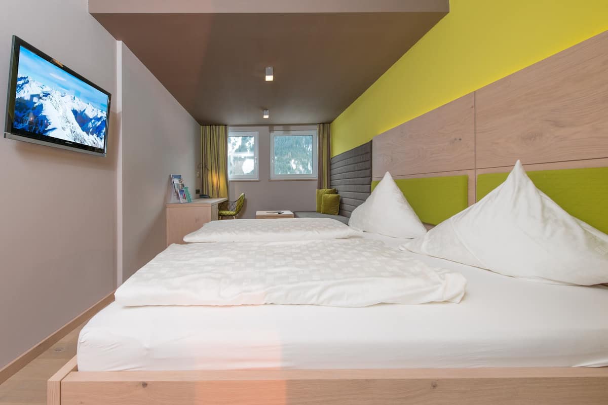 A modern hotel room with a large bed, a flat-screen TV mounted on the wall, and a window providing natural light.