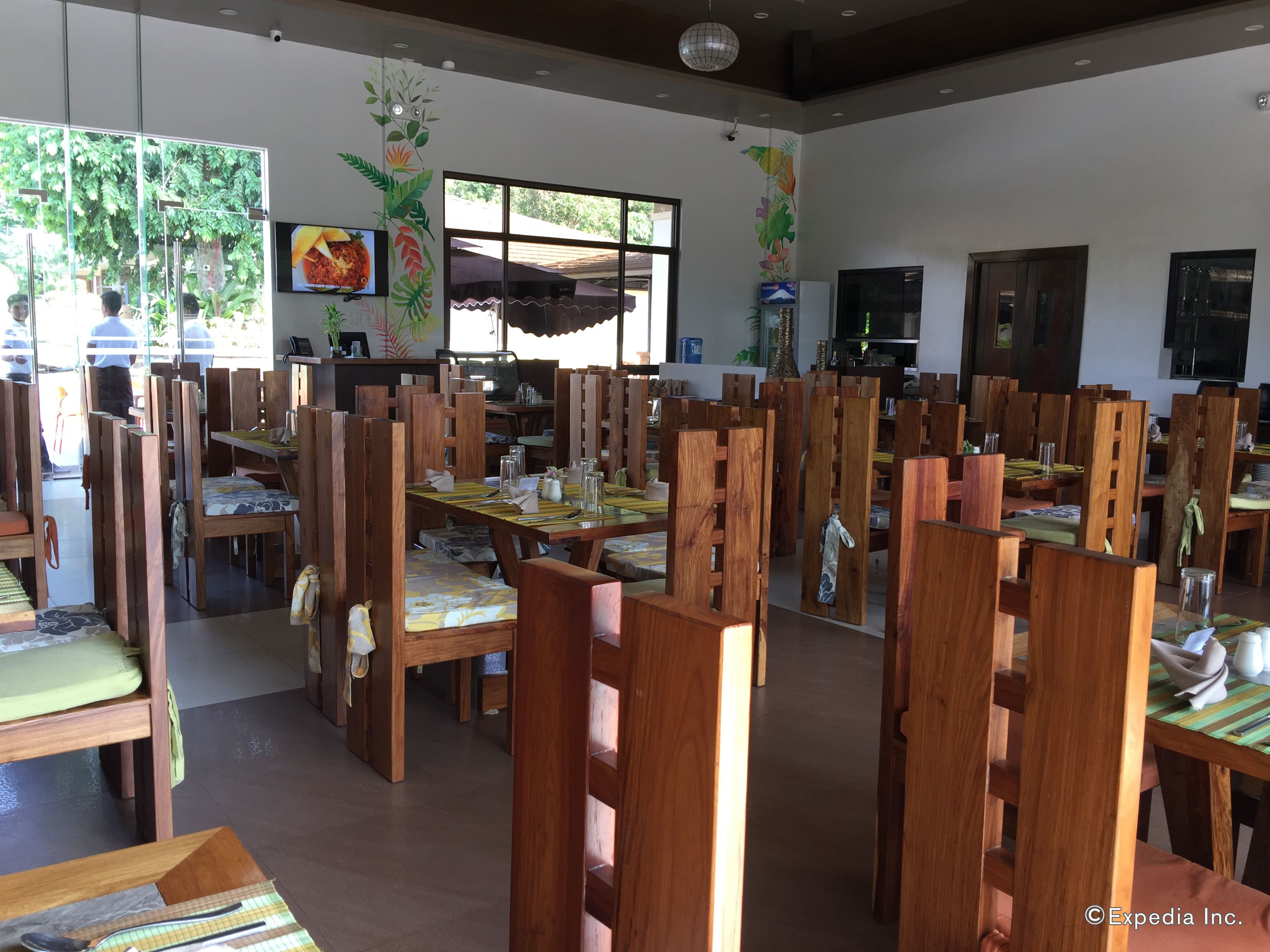 A spacious dining area with wooden tables and chairs, large windows with natural light, and a television mounted on the wall.