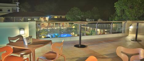 Un área de estar al aire libre con sillas anaranjadas y una mesa de vidrio, con vistas a una piscina y un edificio con balcón.