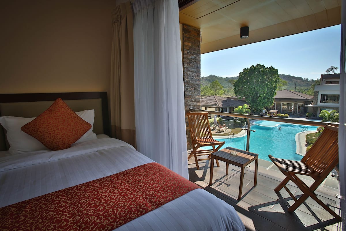 Habitación de hotel con cama, colcha roja estampada y vista a una piscina y zona de estar exterior.