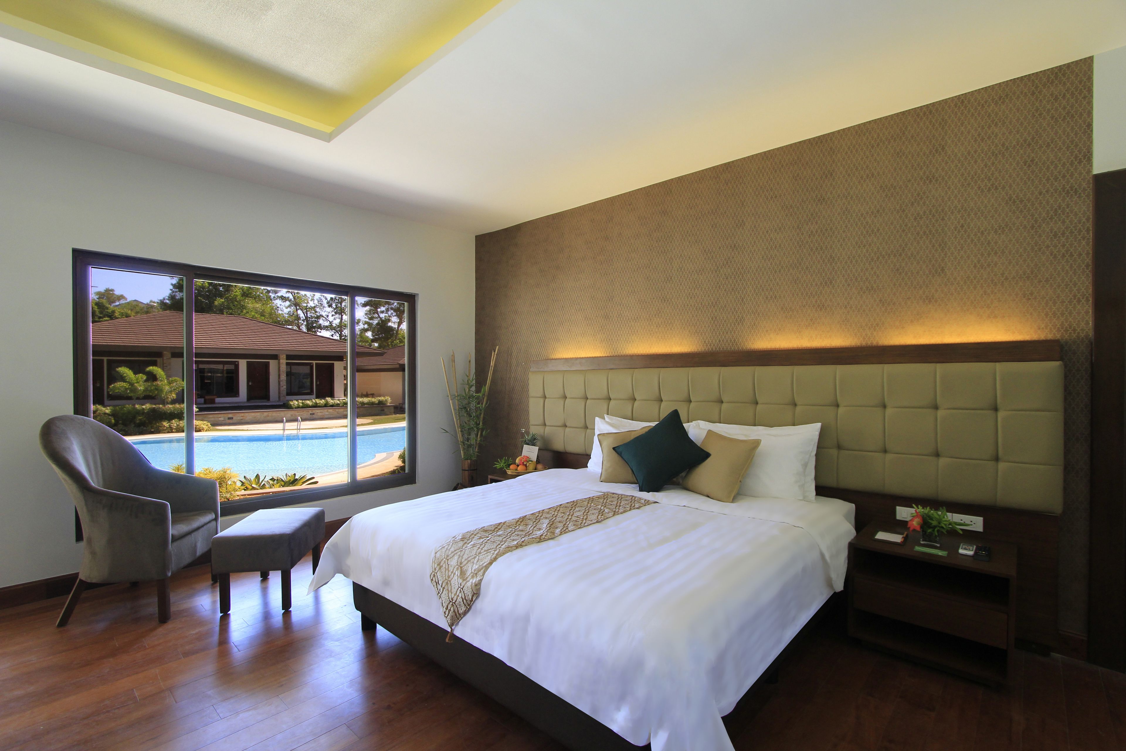 A modern hotel room with a large bed, a sitting area with a chair, a nightstand, and a view of a swimming pool.
