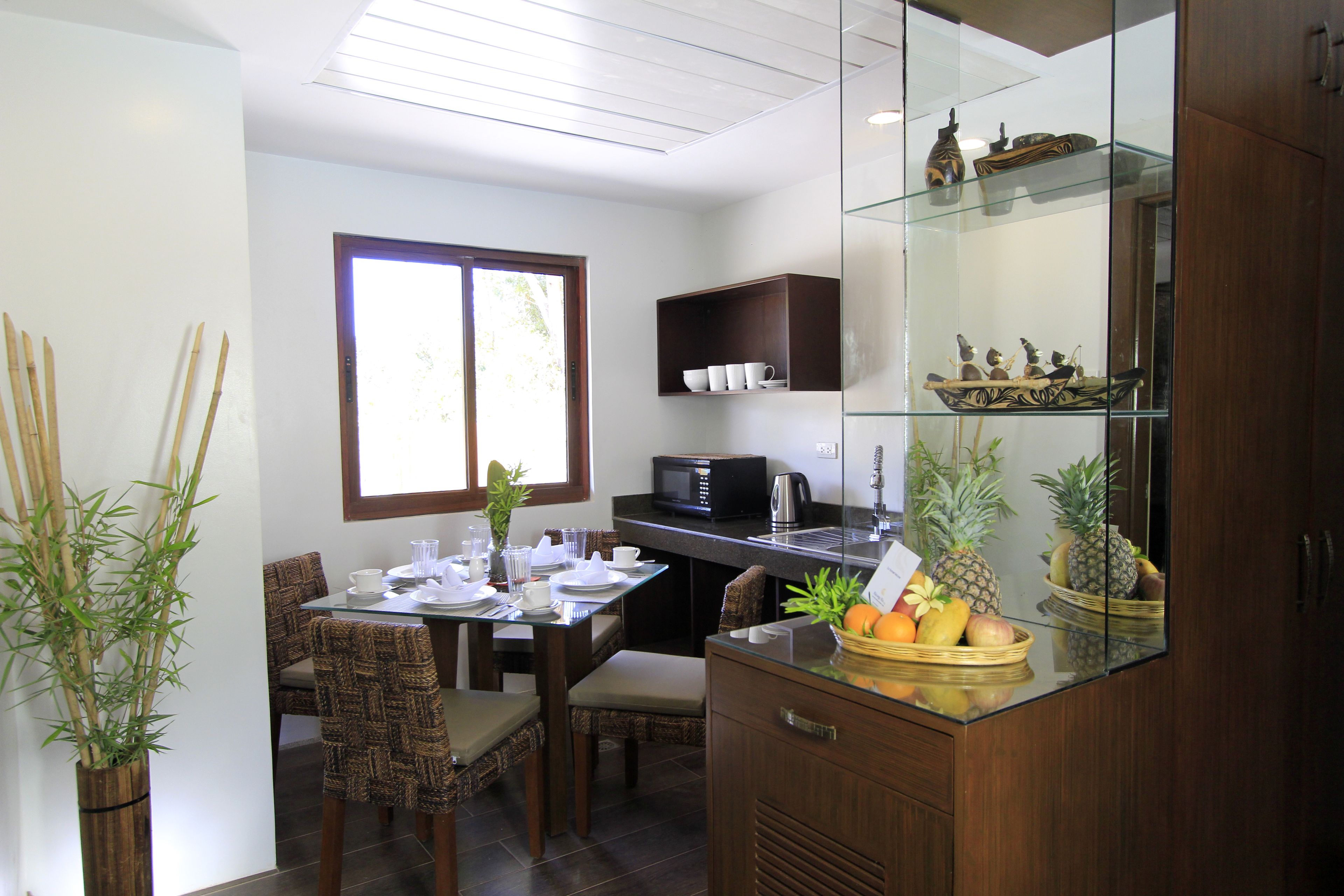 A modern kitchen with a dining area, featuring a glass-top table, wooden chairs, a microwave, and a fruit basket.