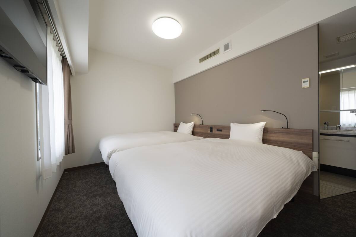 Une chambre d’hôtel avec deux lits, un grand miroir, une fenêtre avec des rideaux et un luminaire au plafond.