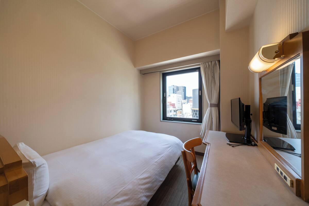 Une chambre d’hôtel avec un lit, une chaise, un bureau avec un ordinateur, une télévision et une fenêtre donnant sur la ville.