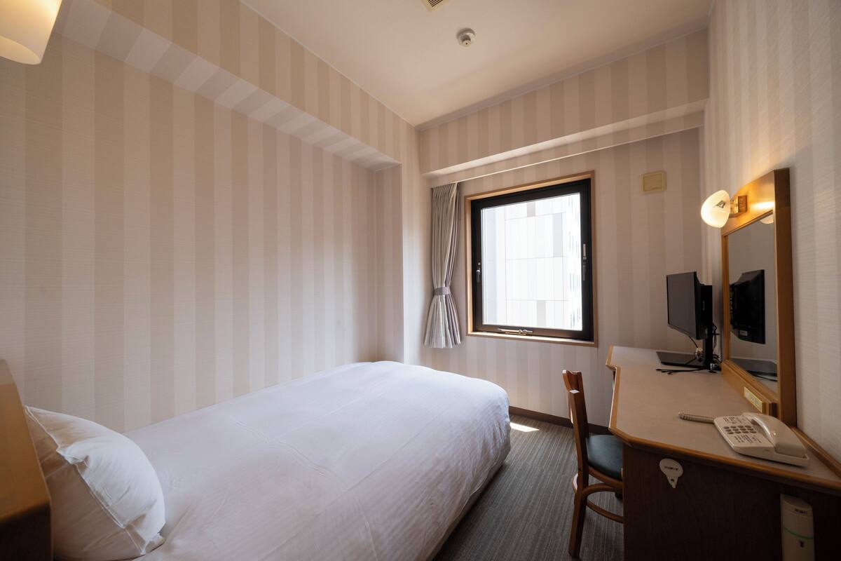 Une chambre d’hôtel comprenant un lit, un bureau avec un téléphone, une chaise, une télévision et une fenêtre avec des rideaux.