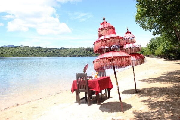 A beachside dining setup with a table and chairs under a red umbrella, overlooking a calm lake with a forested background.