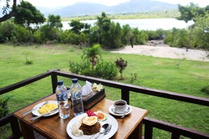 A wooden table with breakfast items, a bottle of water, and a cup of coffee, overlooking a grassy area with trees and a body of water.