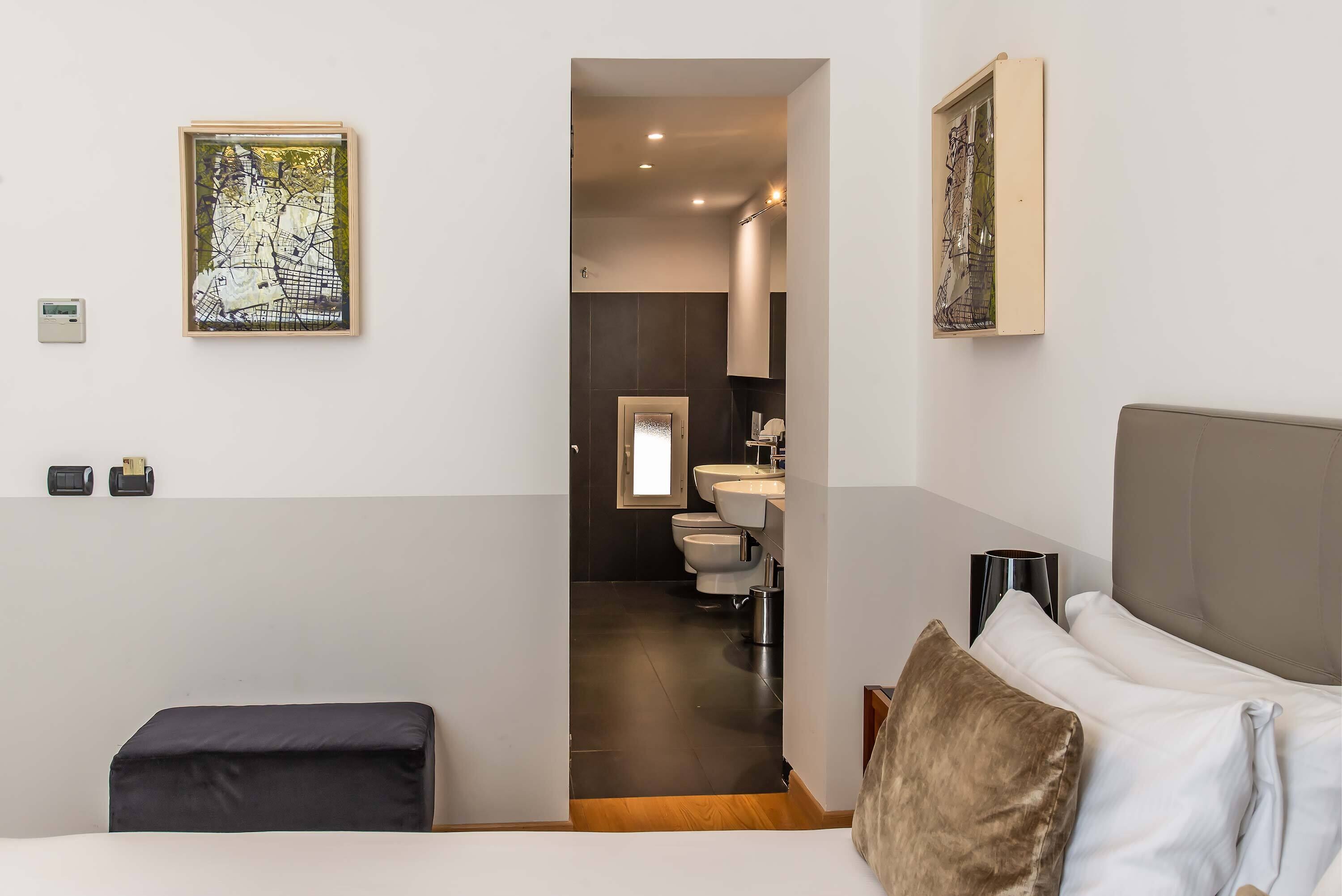 A modern hotel room with a bed, a bedside table, a nightstand, a large window, and a bathroom visible through an open door.