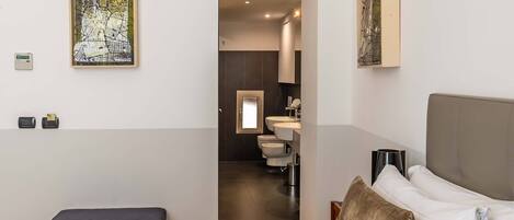 A modern hotel room with a bed, a bedside table, a nightstand, a large window, and a bathroom visible through an open door.