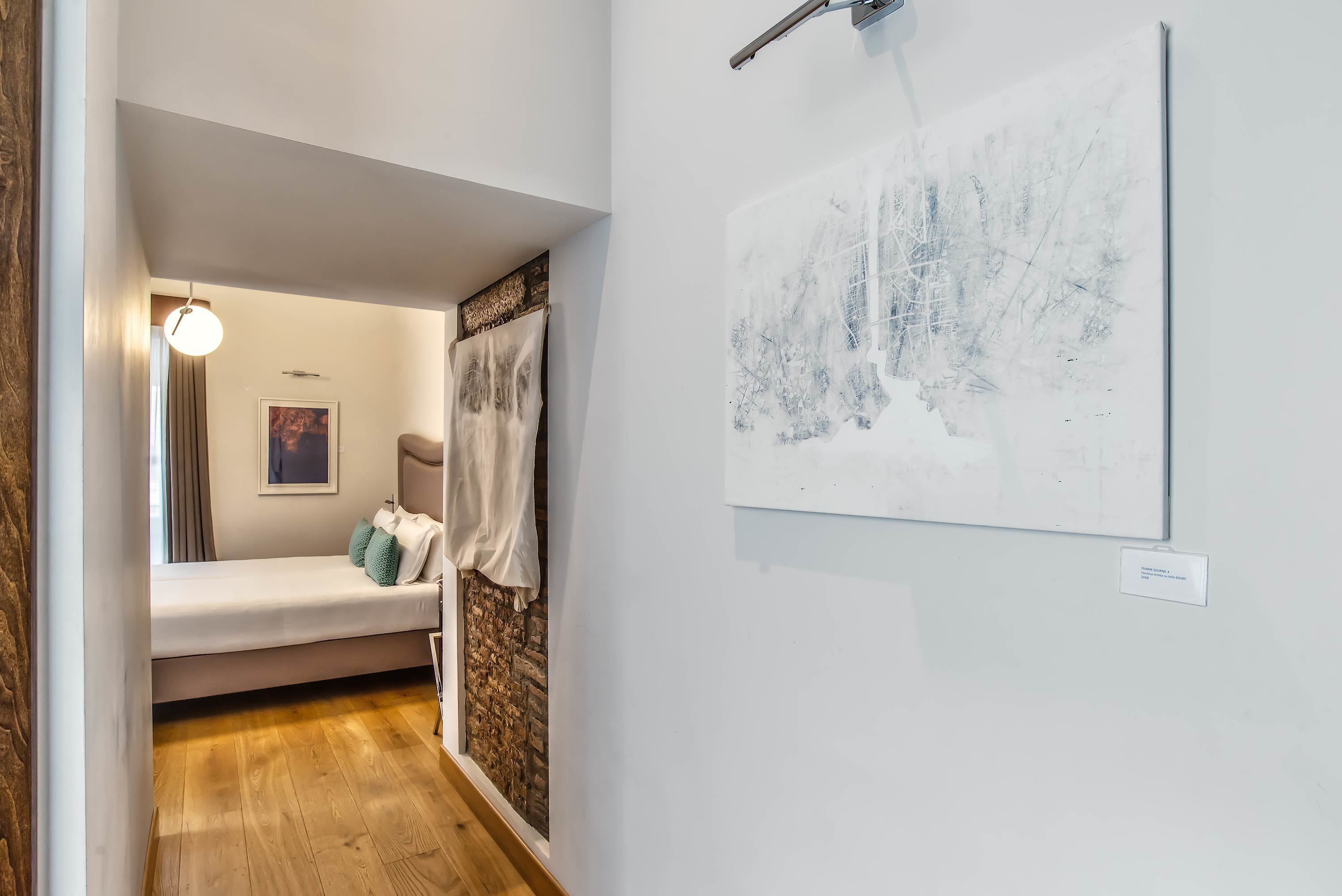 A hallway with a wooden floor, a white wall with a framed artwork, a bed with a white headboard, and a hanging light fixture.
