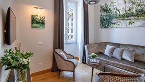 A modern living room with a sofa, two armchairs, a coffee table, a painting, and a view of a building through a window. - Relais Rione Ponte (Rome)