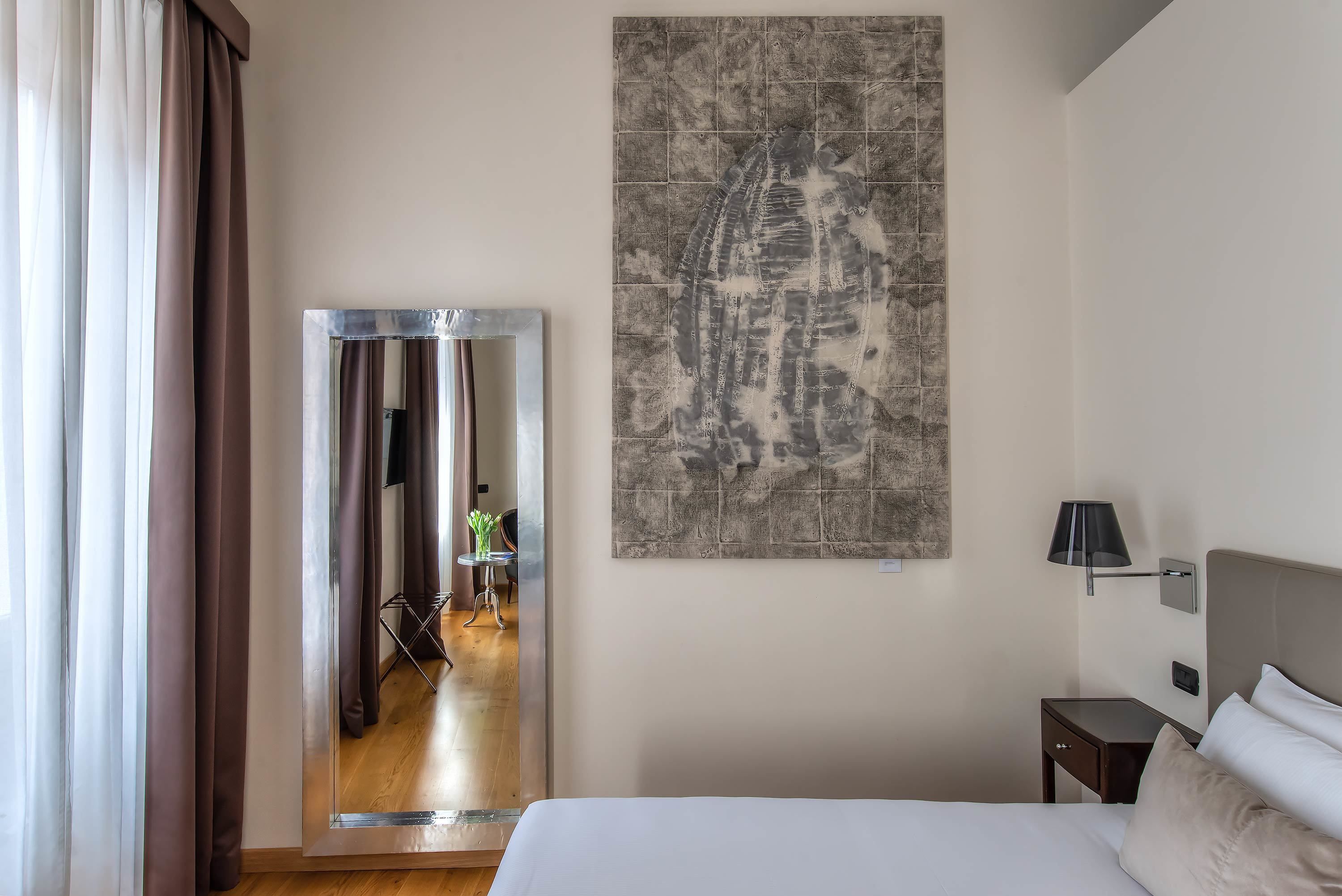 A hotel room with a bed, a nightstand, a lamp, a painting, and a view of a hallway with a person and a chair.
