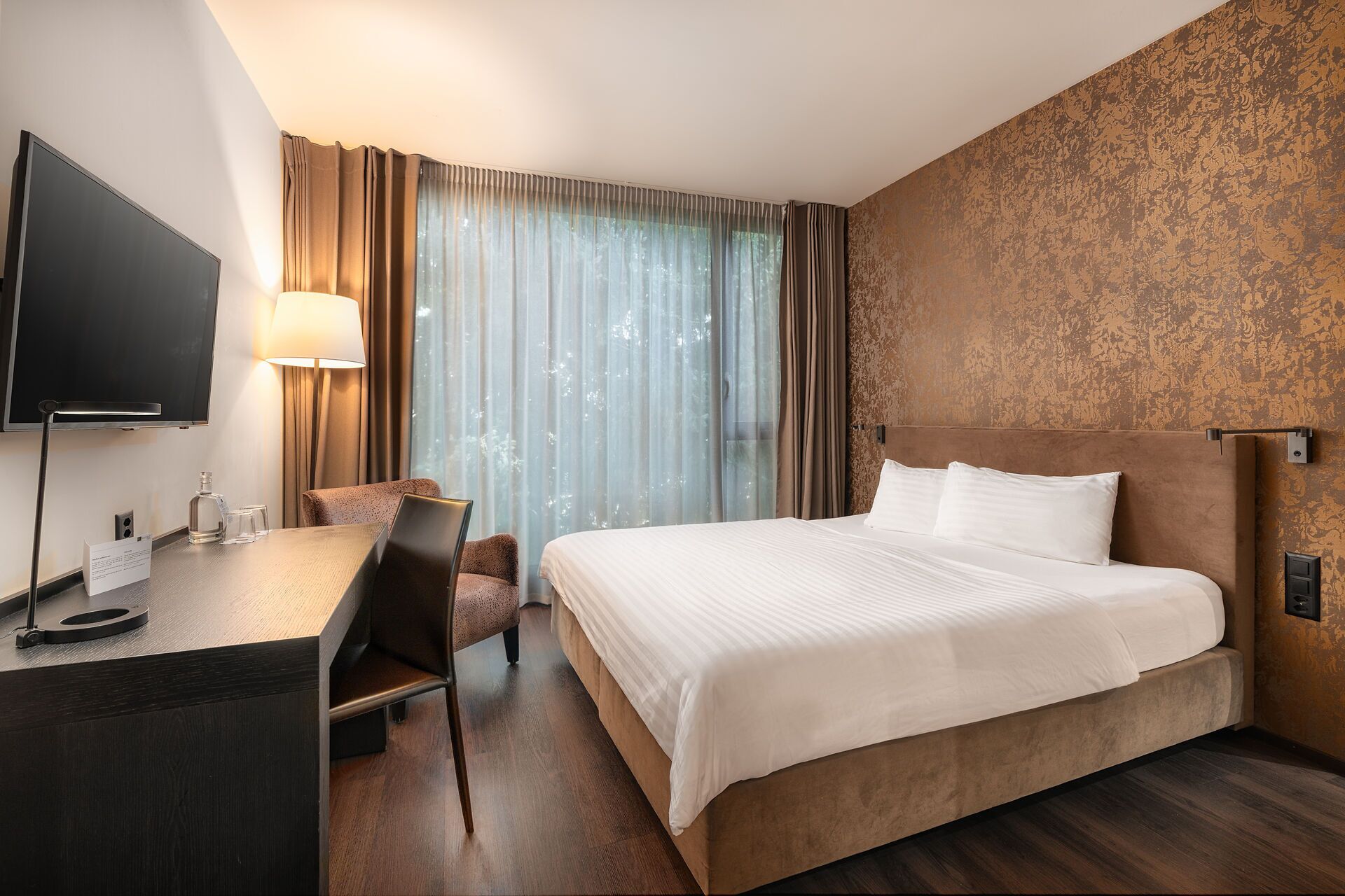 A hotel room with a bed, a desk, a chair, a TV, and a window with curtains.