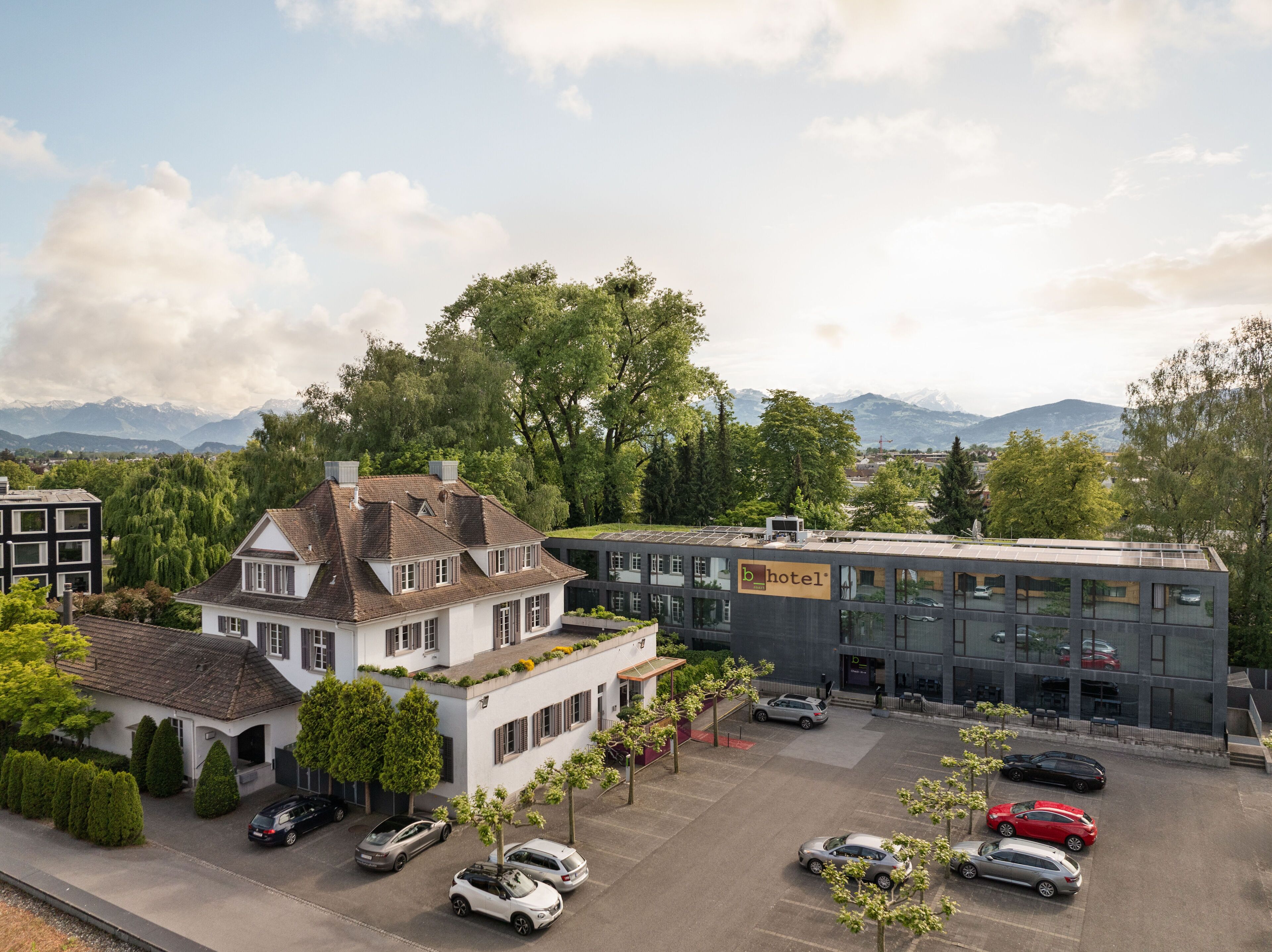 A hotel with a traditional building and a modern extension, surrounded by trees and a parking lot.