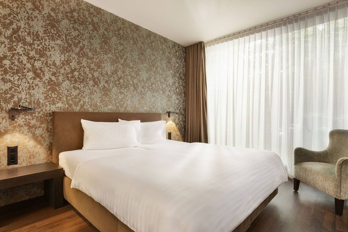A hotel room with a large bed, a nightstand, a chair, and a window with sheer curtains.