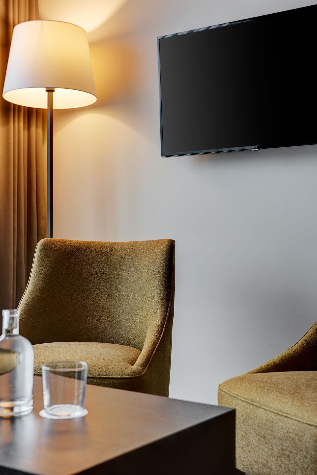 A hotel room with a flat-screen TV mounted on the wall, a brown armchair, a side table with a bottle and glass, and a lamp.