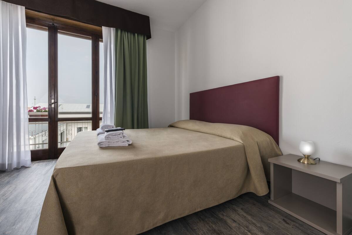 A hotel room with a bed, a nightstand, a window with curtains, and a view of buildings outside.