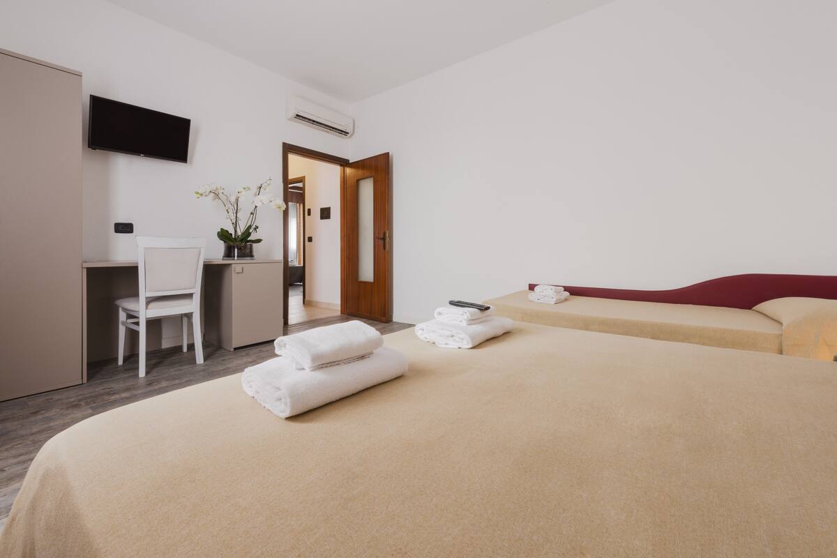 A hotel room with a bed, a desk, a chair, a TV, and a door leading to another room.