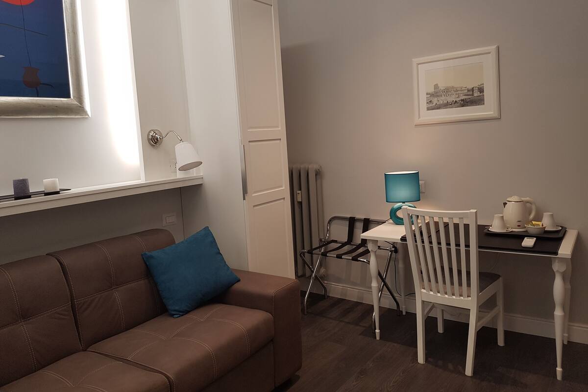 Ein modernes Wohnzimmer mit einem braunen Sofa, einem blauen Kissen, einem weißen Schreibtisch mit Stuhl, einer Lampe und einem gerahmten Bild an der Wand.