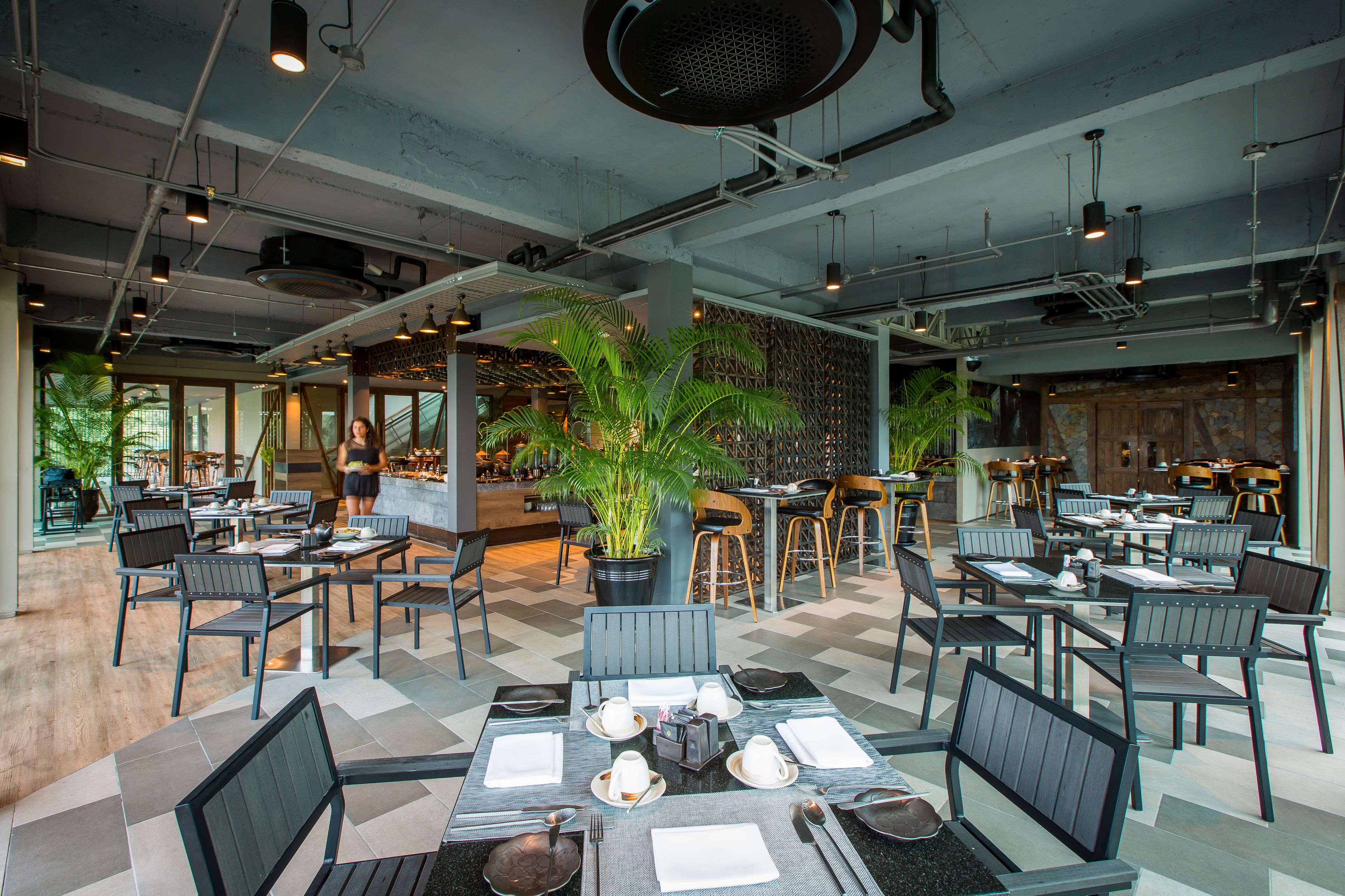A modern restaurant with a mix of indoor and outdoor seating, exposed ceilings, and potted plants.