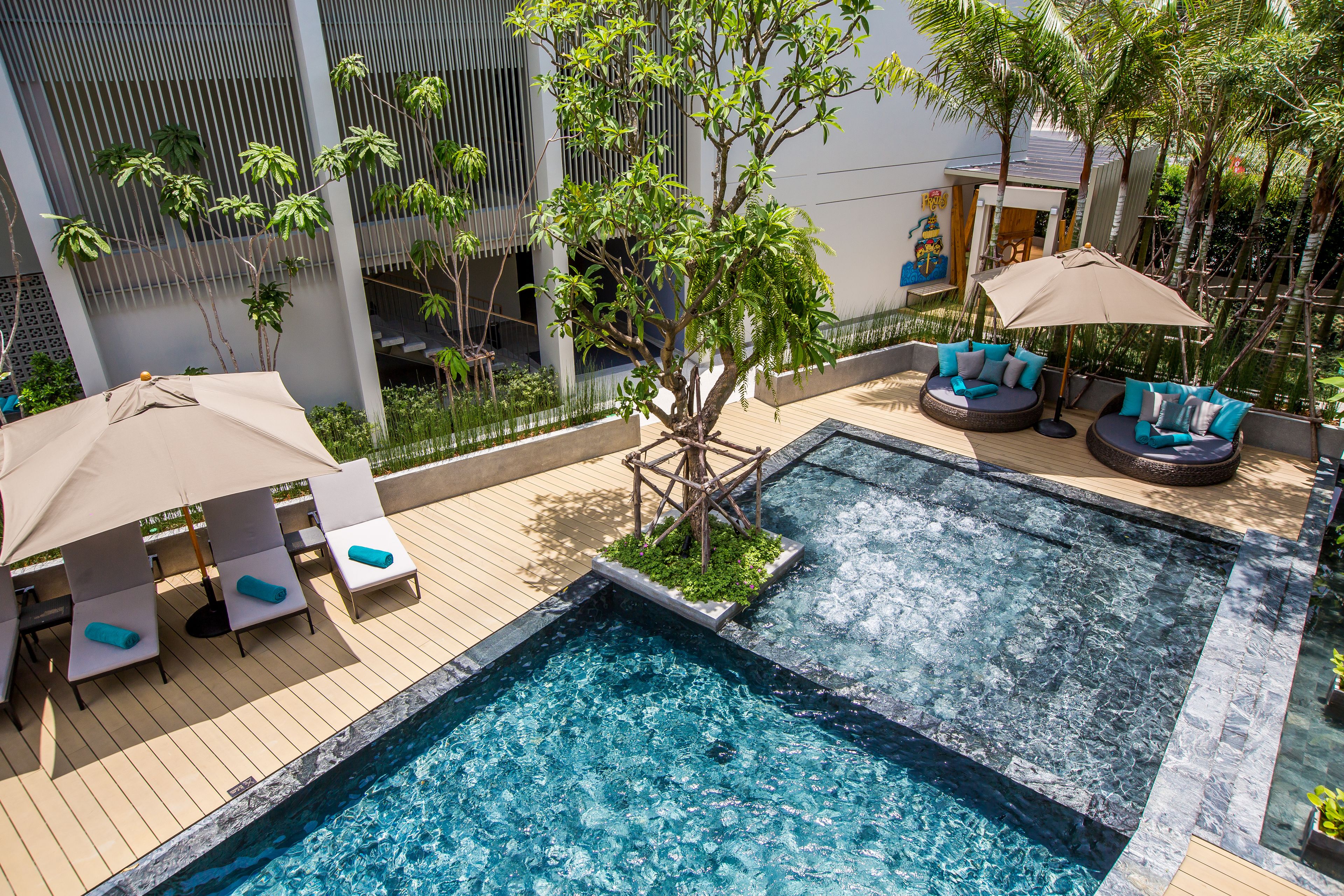 A rooftop pool area with lounge chairs and umbrellas, surrounded by greenery and modern architecture.