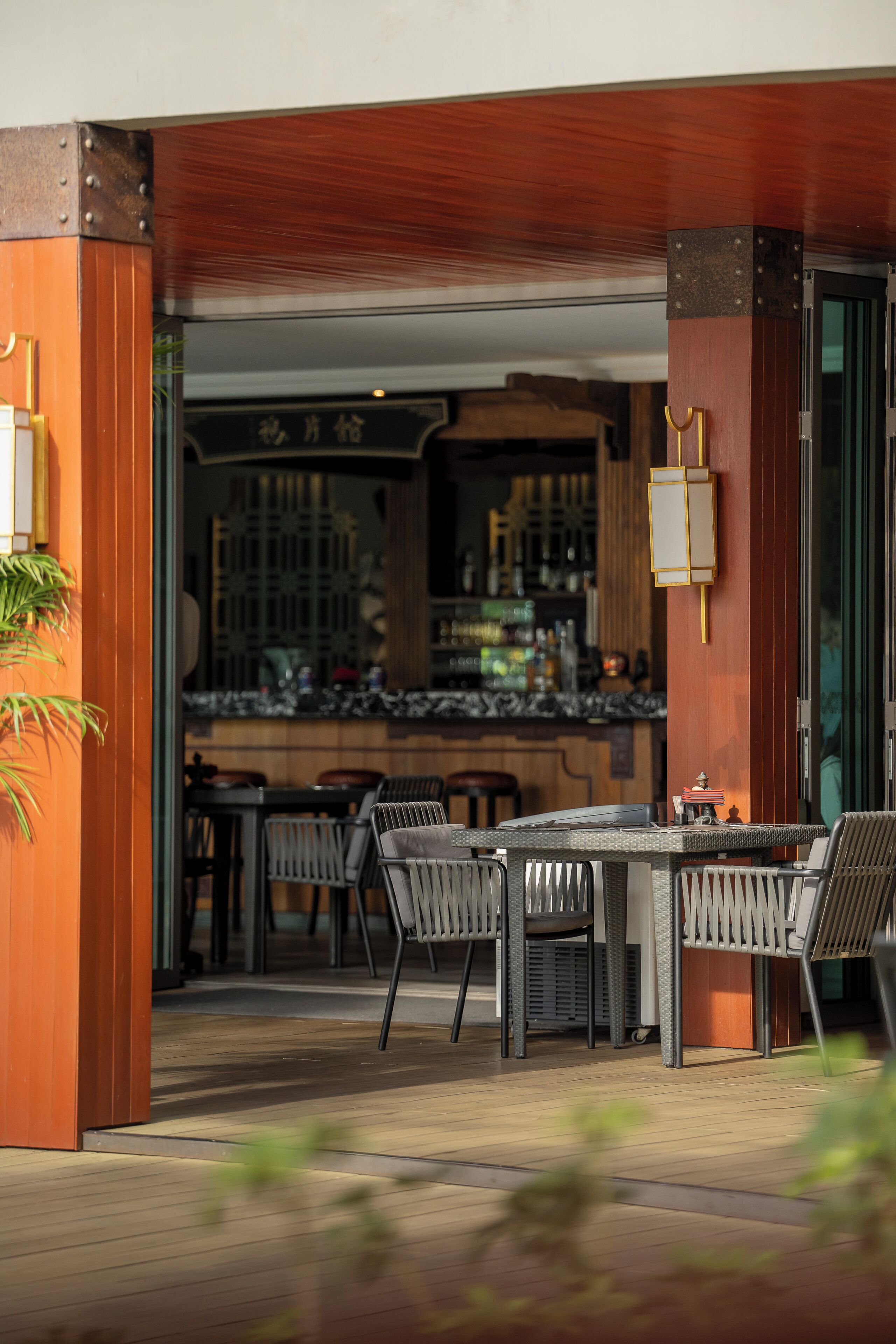 An outdoor seating area with tables and chairs, a bar with shelves of bottles, and a wooden structure with a sign.