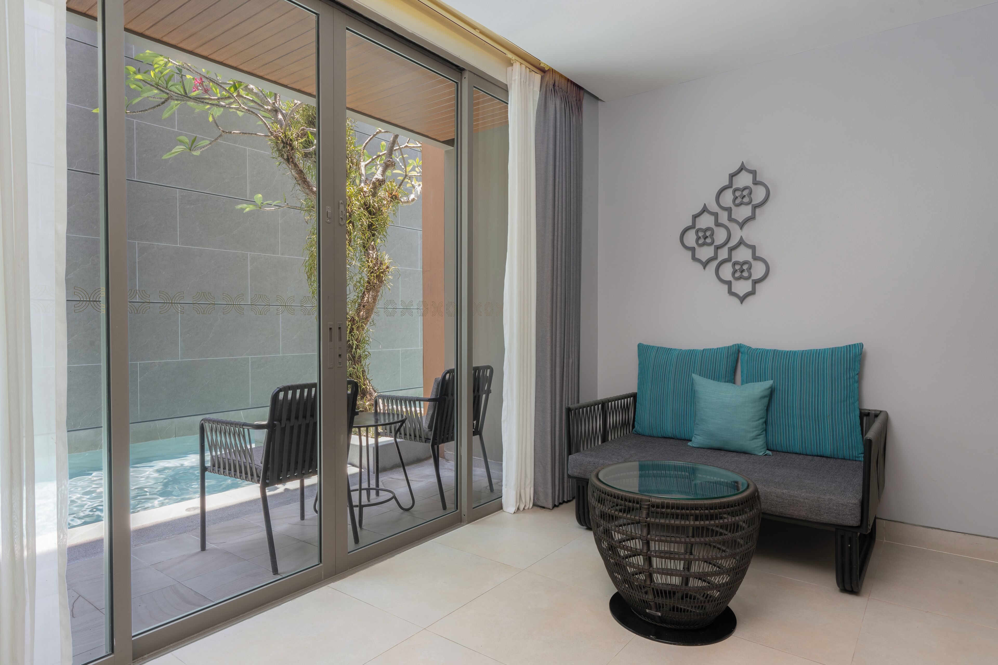 A modern room with a glass door leading to a pool, a sofa with teal cushions, a wicker coffee table, and a decorative wall piece.