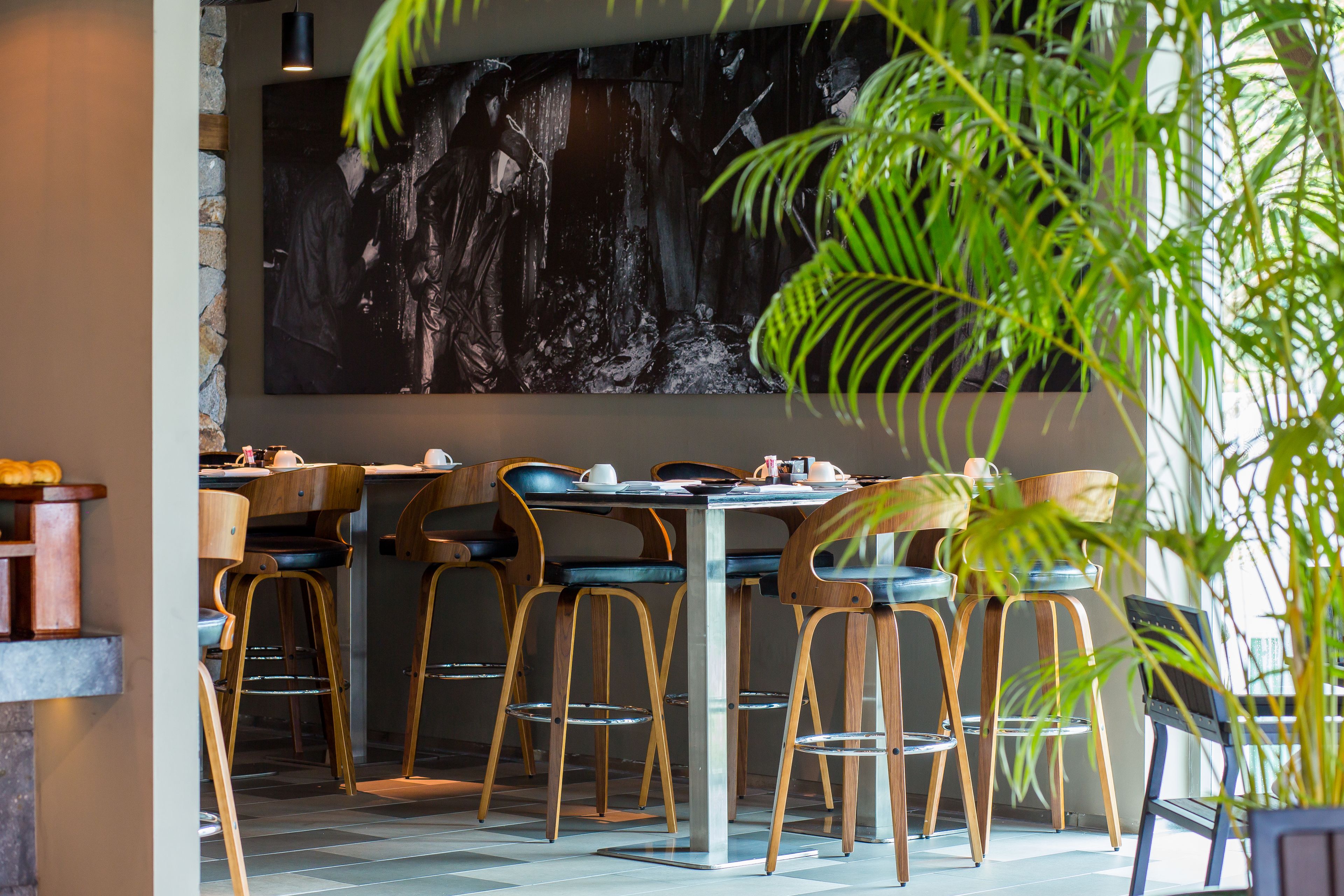 A restaurant interior with wooden chairs and tables, a large abstract painting on the wall, and a potted plant in the foreground.
