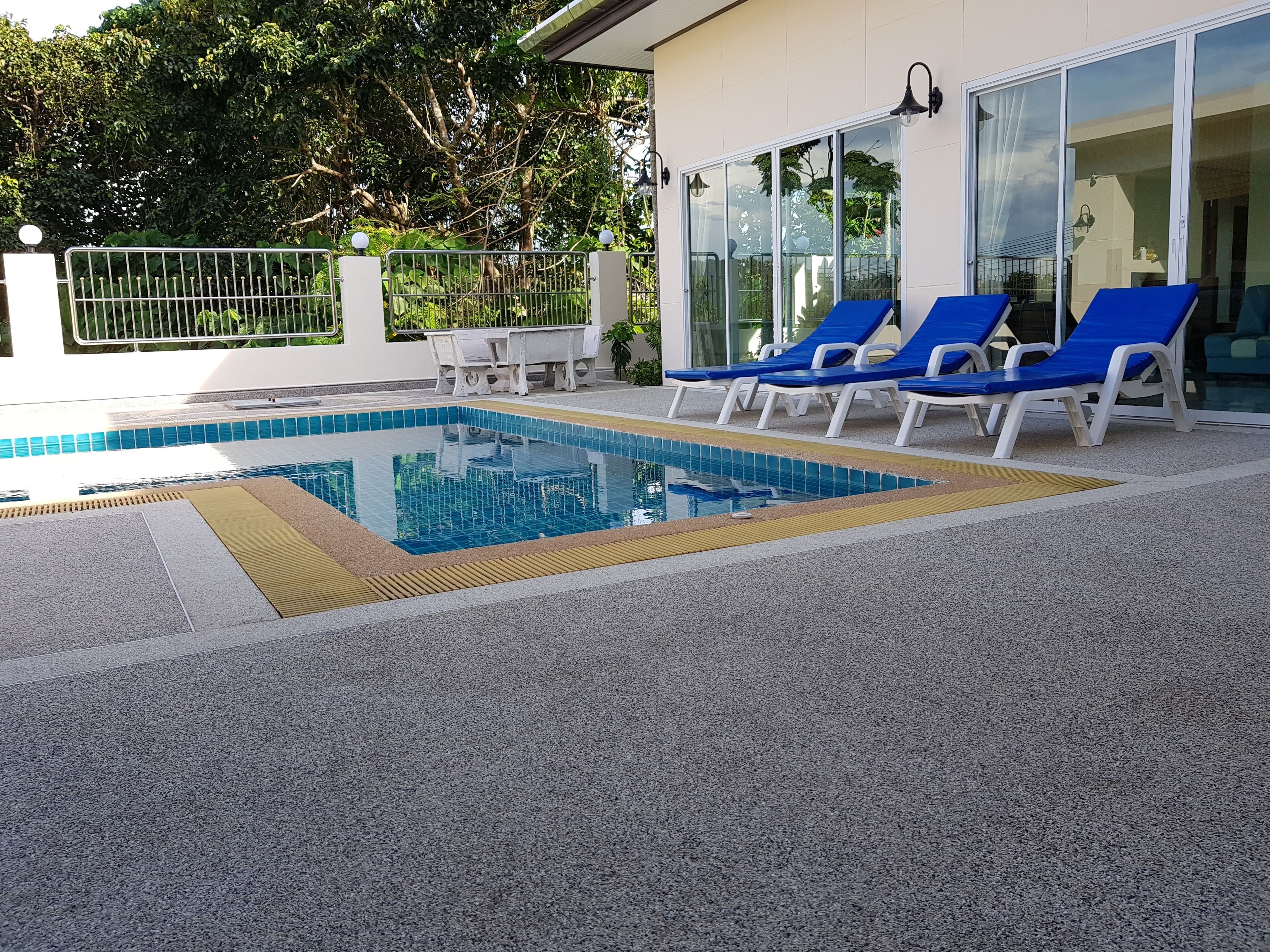 A swimming pool with blue tiles, surrounded by a grey textured deck. There are blue lounge chairs and a white table with chairs.