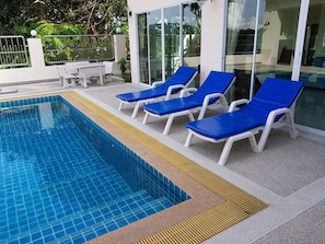 A swimming pool with blue tiles, white lounge chairs, and a glass door leading to a covered area.