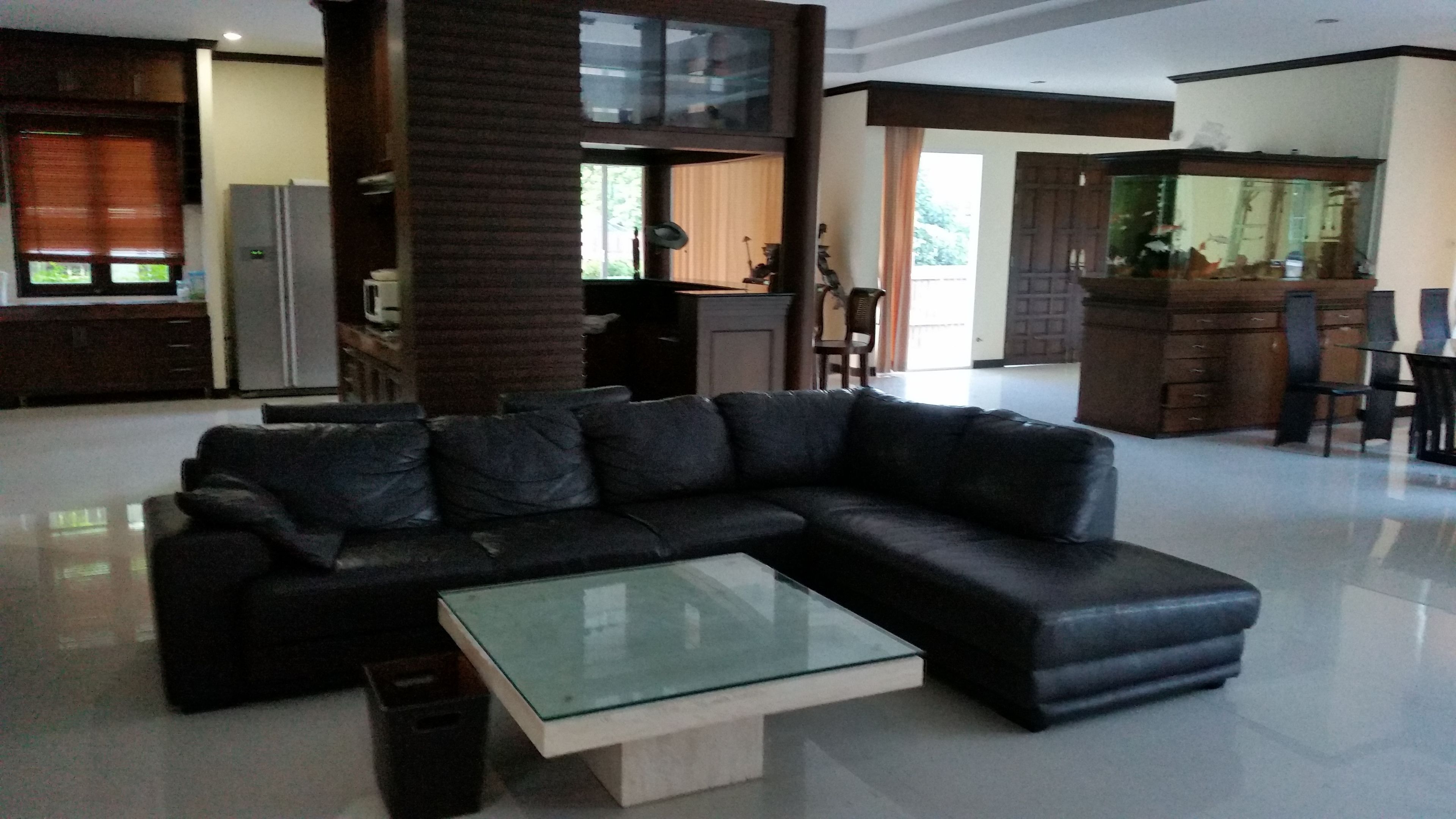 A modern living room with a black leather sectional sofa, a glass coffee table, and a kitchen area in the background.