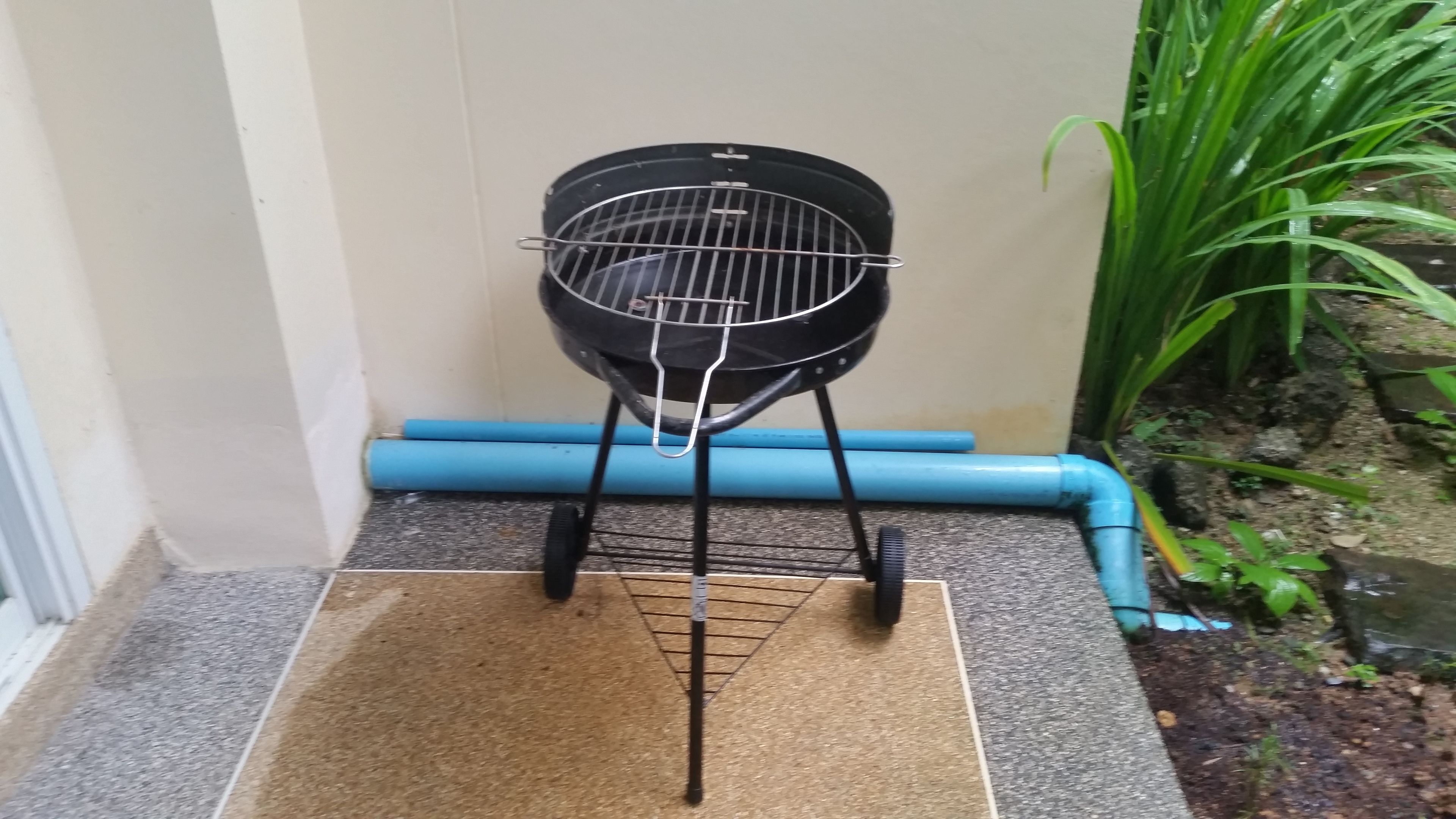 A portable grill with a metal grate and a handle, placed on a mat in a corner with a blue pipe and green plants nearby.