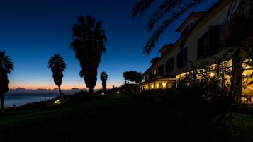 A coastal property with a well-lit building, palm trees, and a view of the ocean at dusk.