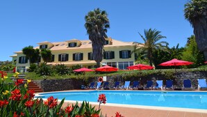 Uma área de piscina com espreguiçadeiras e guarda-sóis, um edifício grande com telhado de azulejos e vegetação tropical.