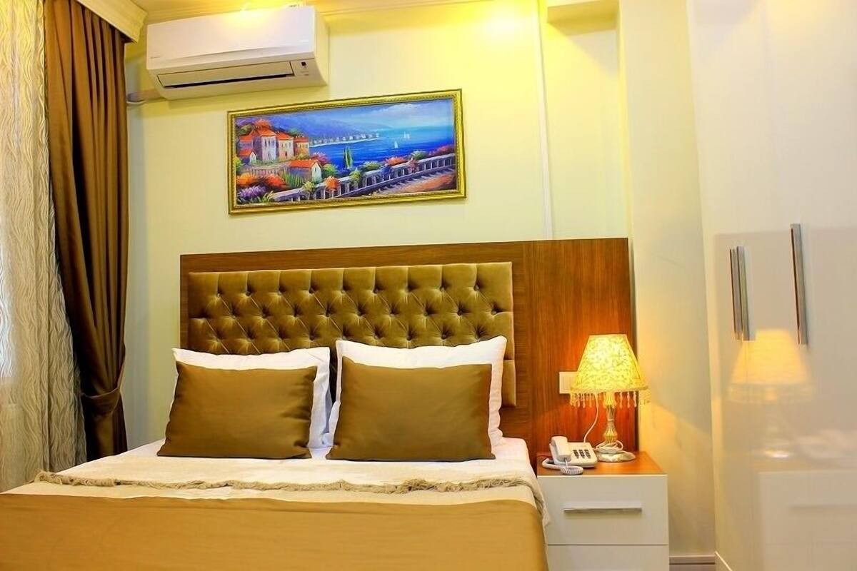 Hotelkamer met een groot bed, een nachtkastje met een lamp, een schilderij van een kustlandschap en een airconditioning.